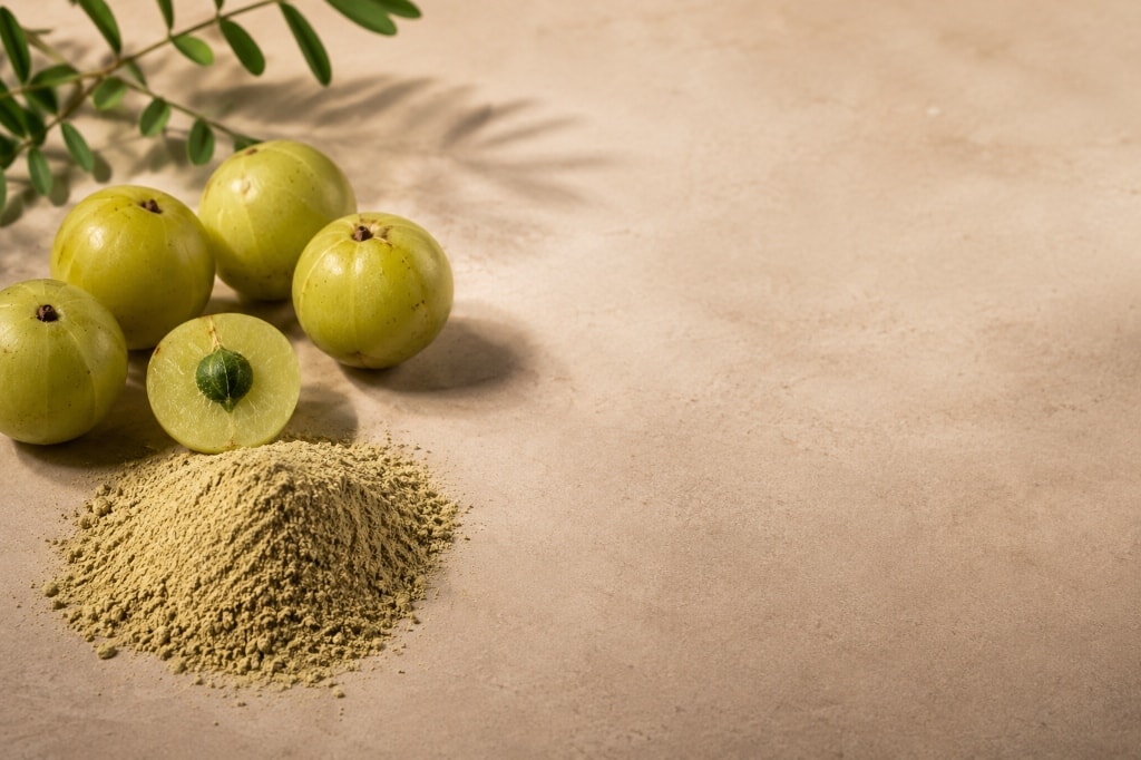 A mound of pale amla powder beside whole and halved green amla fruit.